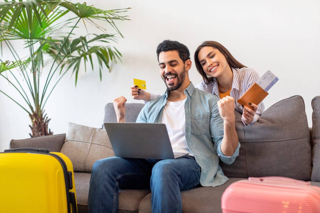 Joyful young tourists couple booking hotel room online, using laptop, woman holding credit card and passports with tickets