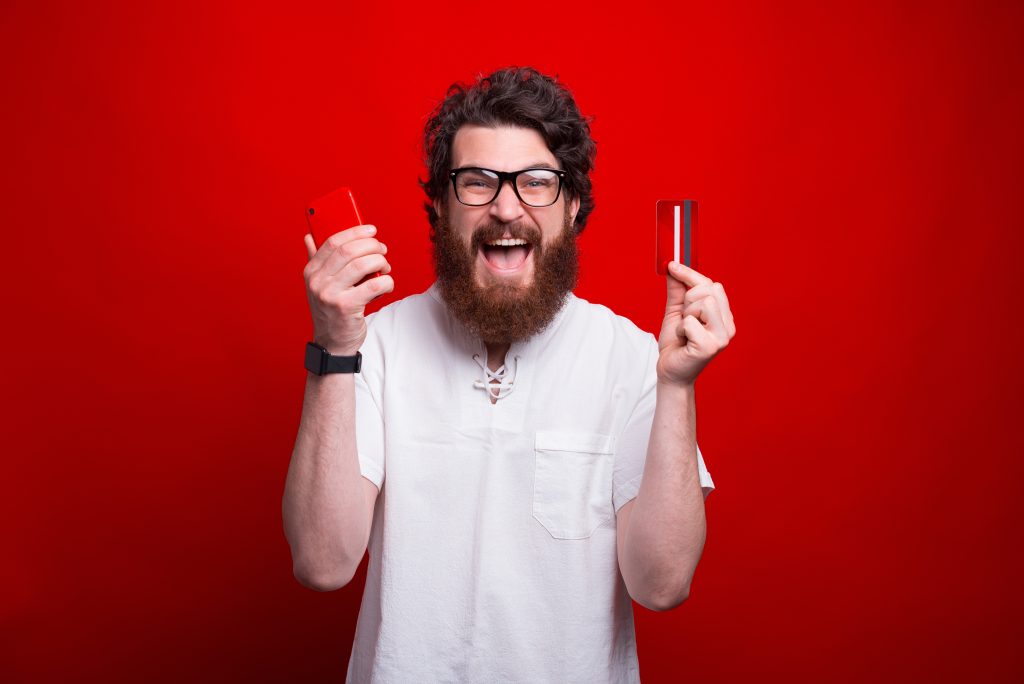 Excited bearded man is holding a phone and credit card over red background