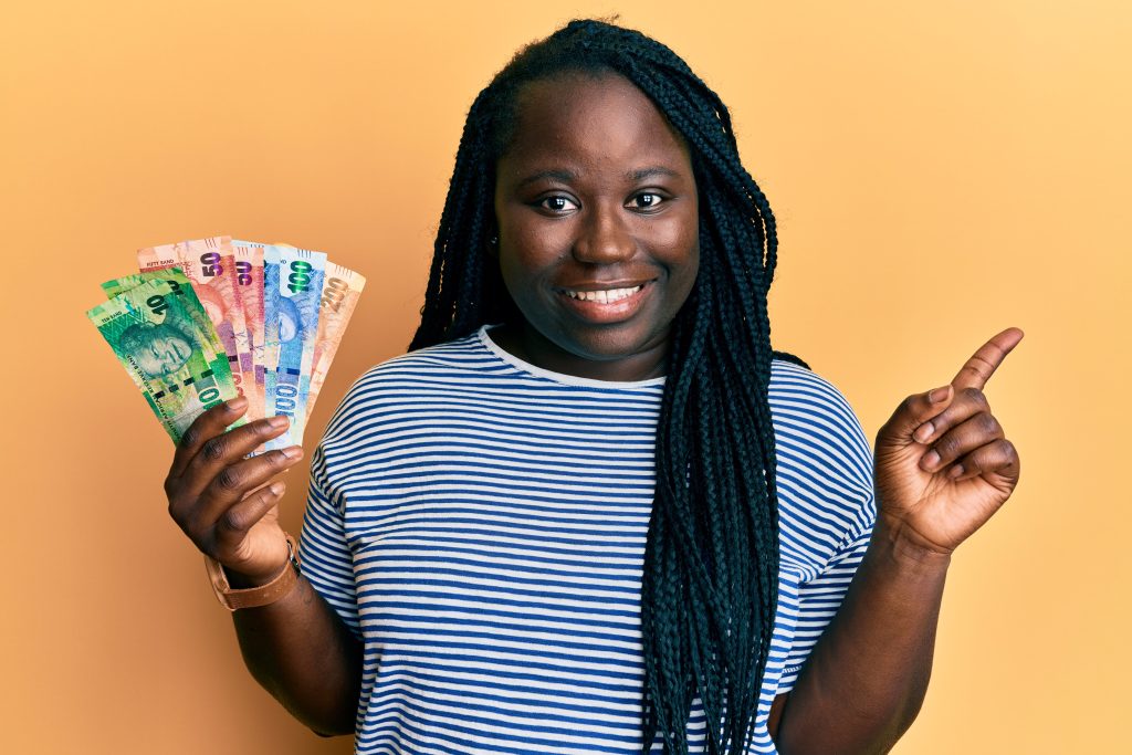 Young black woman with braids holding south african rand banknotes smiling happy pointing with hand and finger to the side