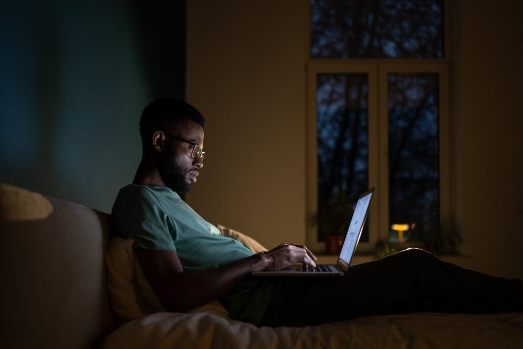 Concentrated African American programmer man lying on bed work on laptop at night.