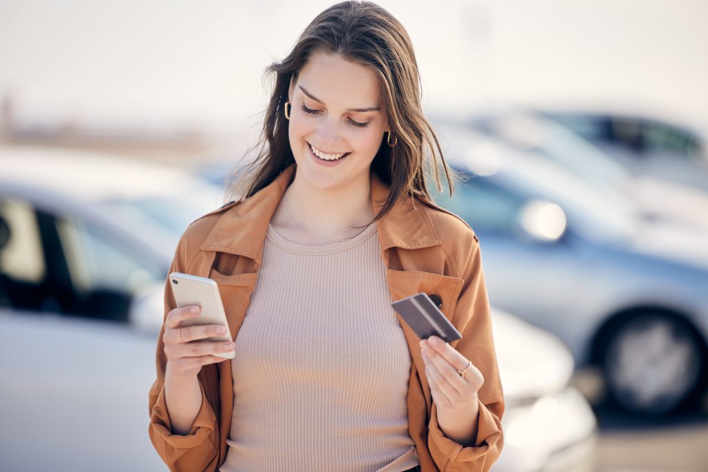 Car shopping for the day. Shot of a young woman using her card and phone outside.