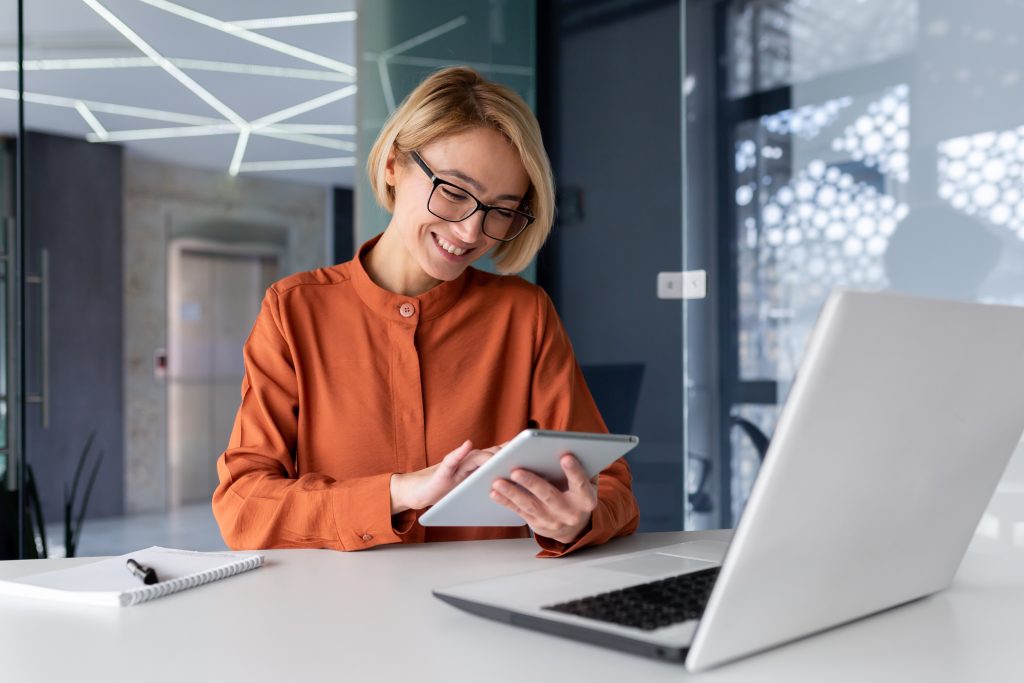 Young successful businesswoman at workplace is satisfied with results of achievement at work smiling and using tablet computer, blonde programmer is testing new software in application