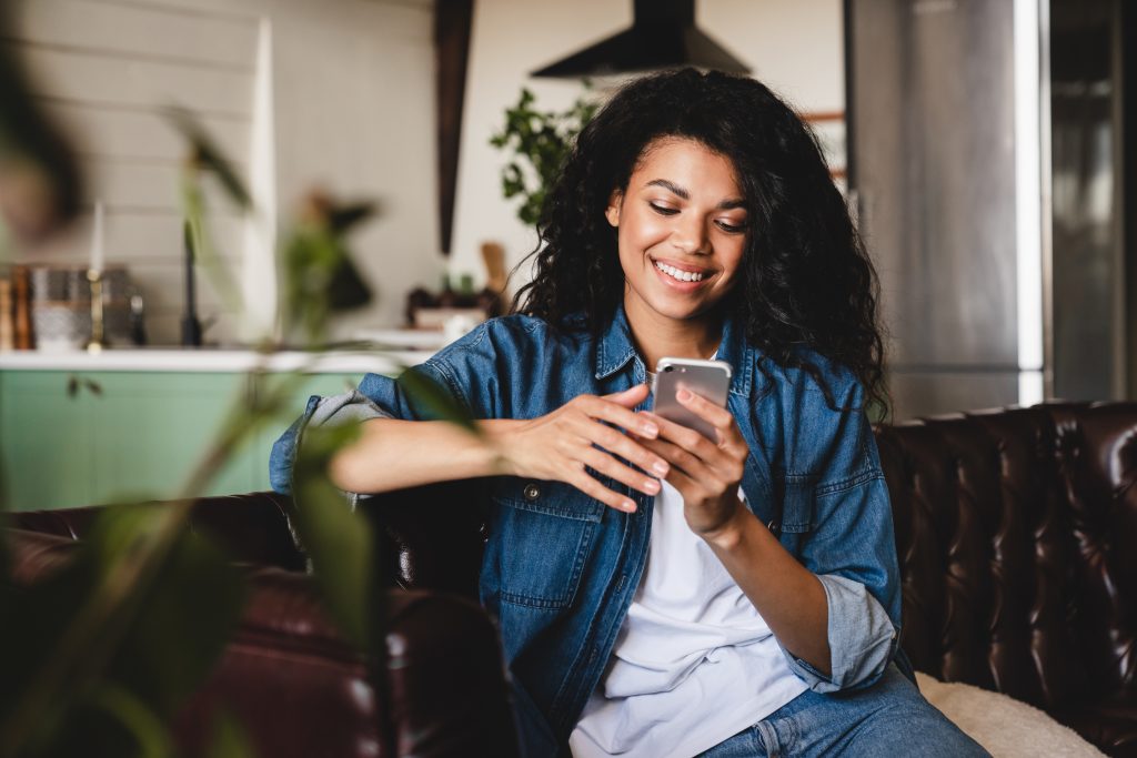 Relaxed young african woman texting on her phone in the modern house.Smiling african american woman using smartphone at home, messaging or browsing social networks while relaxing on couch