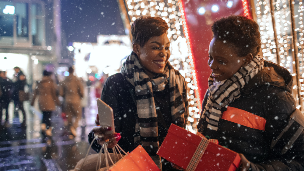 two black women holding shopping bags and smiling in the snow while Christmas shopping