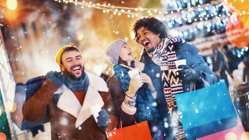 three people laughing in the snow while holding Christmas shopping bags