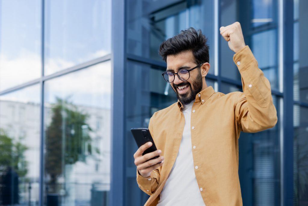 Young successful hispanic man in casual clothes outside office building received online win notification, man celebrating victory triumph, using app on phone, good news in businessman